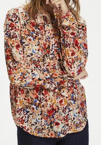 Mujer que lleva una blusa floral de manga larga y con botones, con patrones en rojo, azul, naranja y beige, combinada con pantalones de color oscuro.