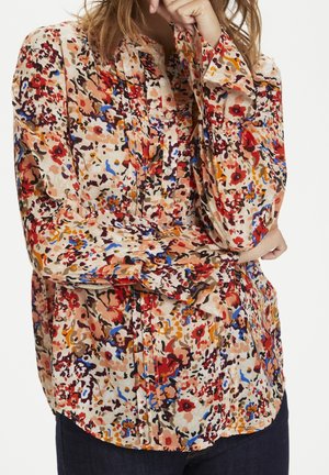 Femme portant une blouse florale à manches longues et boutonnée, avec des motifs rouges, bleus, orange et beige, associée à un pantalon de couleur sombre.