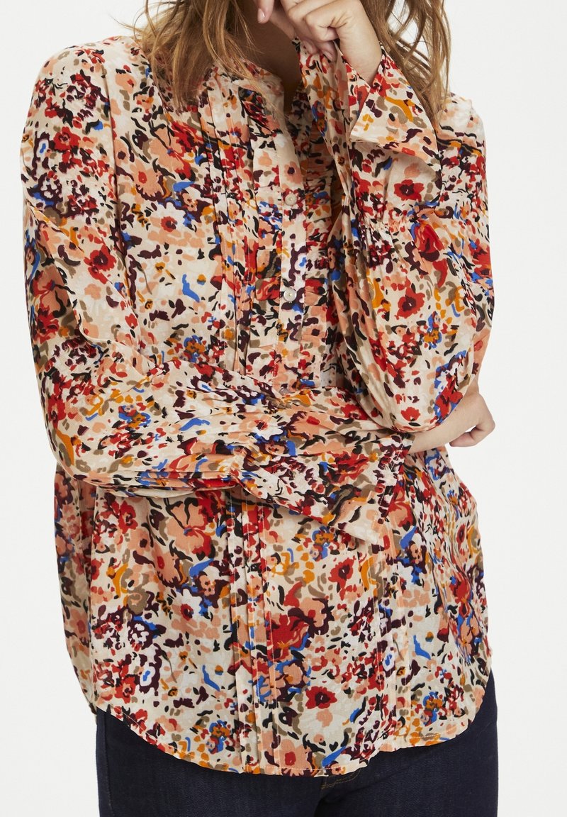 Mujer que lleva una blusa floral de manga larga y con botones, con patrones en rojo, azul, naranja y beige, combinada con pantalones de color oscuro.