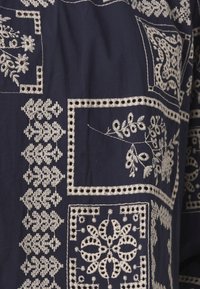Le tissu marine présente une broderie blanche complexe avec des motifs géométriques et floraux. Comprend des accents bordés et divers designs de points.