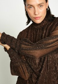 Ruda tekstūruota bliuskutė su aukštu kaklu ir gausiomis rankovėmis. Medžiaga turi metalinį siūlų akcentą ir raukšlėtus manžetus.