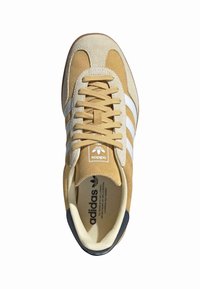 Tan spalvos nubuko sneakeriai su baltais dryžiais, apvaliu pirštu ir plokščiomis raištelėmis. Ant liežuvėlio yra logotipas, o kontrastinga tamsi kulno akcentacija.