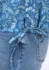 Blaue Blumenbluse mit grünen Akzenten; verfügt über einen seitlichen Knoten und einen Schlitz. Kombiniert mit hellblauen Jeans, die einen strukturierten Stoff zeigen.