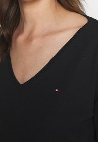 Czarny, prążkowany top z dekoltem w serek z subtelną naszywką logo w czerwieni, bieli i niebieskim. Materiał wydaje się miękki i ma dopasowany krój.