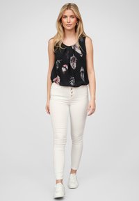 Svart ermeløs topp med fjærtrykk, parret med hvite skinny jeans med knappfront og hvite joggesko.