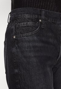 Mustat farkut, joissa on lähikuva vyötäröstä, taskuista ja metallinappista, sekä hillitty tekstuuripinta.