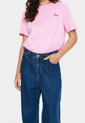 Femme portant un t-shirt rose pâle avec le mot "love" et un petit cœur, associé à un jean en denim bleu foncé taille haute, main dans la poche.