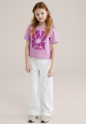 Lila katoenen T-shirt met een grote paarse bloemenafbeelding en tekst, gecombineerd met witte wijde broek en lichtgekleurde sneakers.