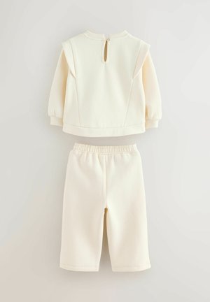 Set a due pezzi color crema chiaro composto da una maglietta a maniche lunghe con chiusura a zip sul retro e pantaloni corti coordinati con vita elasticizzata.