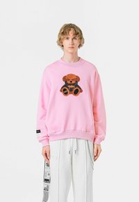 Sudadera rosa claro con un diseño de oso peludo en la parte frontal, con acentos en negro. Puños y dobladillo acanalados, ajuste relajado.
