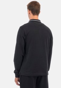 Svart långärmad polotröja med platt krage, vita accentstrimmor och sidofickor. Tillverkad av ett mjukt och slätt tyg.