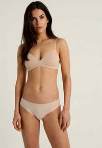 Woman with long dark hair wearing nude-colored bra and matching briefs, standing against plain light background, facing forward.
