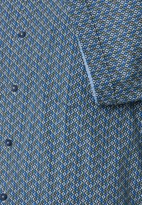Shirt met een geometrisch patroon in tinten blauw en groen, gemaakt van textuurstof met zwarte knopen en een contrasterende kraag.