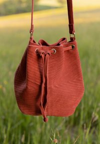 Kabelka typu bucket v rezavé semišové textuře s prošívaným vzorem, stahovací šňůrkou a kovovými průvleky. Nastavitelný popruh pro všestranné nošení.