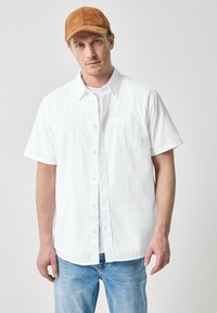 Witte overhemd met korte mouwen en knopen aan de voorkant, met een zak, gedragen over een eenvoudig wit T-shirt, gecombineerd met lichtblauwe spijkerbroek en een bruine pet.