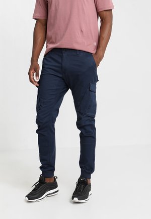 Mann trägt marineblaue Cargo-Jogginghosen, schwarz-weiße Sneaker und ein staubrosa kurzärmeliges T-Shirt, steht mit den Händen in den Taschen.
