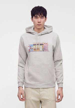 Joven con sudadera con capucha gris claro con ilustración colorida de una cafetería callejera en el pecho y pantalones beige sobre fondo liso.