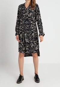 Mustanmutsu pitkähihaisella mekolla, jossa on valkoinen ja beige kukkakuosi, V-pääntie, polvipituus ja sivuraot, yhdistettynä mustiin nauhakengiin.