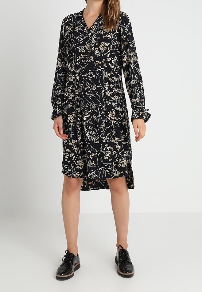 Mustanmutsu pitkähihaisella mekolla, jossa on valkoinen ja beige kukkakuosi, V-pääntie, polvipituus ja sivuraot, yhdistettynä mustiin nauhakengiin.