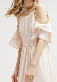 Vestido de color rosa pálido con diseño de hombros descubiertos, mangas cortas con volantes, cuello adornado en dorado y cintura fruncida para una silueta fluida.