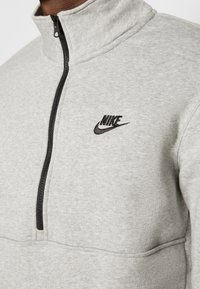 Zbliżenie na jasnoszarą bluzę Nike z krótkim suwakiem z przodu, z czarnym zamkiem błyskawicznym i wyhaftowanym czarnym logo Nike na piersi.