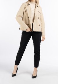 Veste beige à double boutonnage en tissu léger, associée à un pantalon noir slim et des escarpins noirs fermés, sur fond blanc.