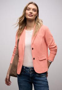 Femme aux longs cheveux blonds portant un blazer corail texturé, une chemise blanche, un jean bleu et un sac à bandoulière marron, sur un fond uni.