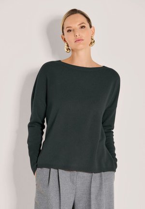 Frau in einem dunkelgrünen langärmeligen Pullover, grauen plissierten Hosen und großen goldenen Creolen, steht vor einem hellbeigen Hintergrund.