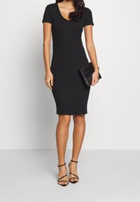 Schwarzes, geripptes Bodycon-Kleid mit kurzen Ärmeln, V-Ausschnitt und taillierter Silhouette, kombiniert mit einer strukturierten schwarzen Clutch und Riemchen-Heels.