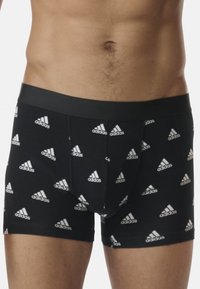 Boxer in cotone elasticizzato neri con un elastico liscio, caratterizzati da un motivo ripetuto del logo Adidas bianco su tutta la superficie.