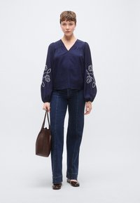 Μπλε μπλούζα με κέντημα λουλουδιών στα μανίκια μπαλόνι, συνδυασμένη με σκούρο τζιν. Το μοντέλο κρατά καφέ υφαντή τσάντα χειρός. Απλά μπαλέτα παπούτσια.