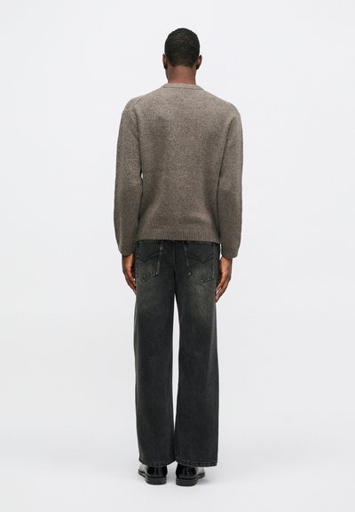 Vīrietis, pagriezies ar muguru, ģērbies pelēkā adītā džemperī, melnās platās biksēs un melnās kurpēs, uz vienkārša balta fona.
