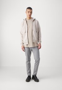 Sudadera cremallera beige sobre una camiseta de color beige claro, combinada con pantalones de cuadros grises y zapatos negros con acentos amarillos.