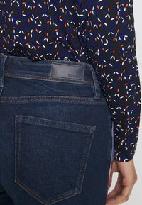 Džínové kalhoty tmavě modré barvy s oranžovým prošitím. Mají logo na pásku a zadní kapsu s přímým švem.