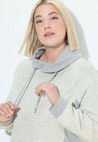 Donna con capelli biondi a caschetto che indossa un felpa grigia chiara, mentre tiene i lacci del cappuccio, su uno sfondo bianco uniforme.