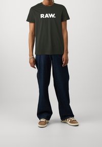 Mørkegrøn bomuld t-shirt med hvid "RAW." print, kombineret med vidde bukser i mørk denim og brune sneakers. Enkel, afslappet outfit.