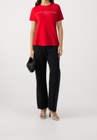 Rotes Baumwoll-T-Shirt mit Druck "TOMMY HILFIGER EST. 1985", kombiniert mit hoch taillierten schwarzen weit geschnittenen Jeans und weißen Riemchenabsätzen.
