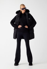 Crna puffer prsluk s umjetnom krznom na kapuljači, preko crnog džempera s pletenim uzorkom. Elegantne crne flared hlače i masivne crne cipele upotpunjuju izgled.