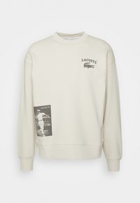 Sweat Lacoste écru col rond, logo Lacoste noir et texte sur la poitrine, graphique joueur de tennis noir et blanc en bas à gauche.