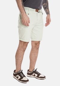 Pantaloni corti verde chiaro con una vestibilità rilassata, risvolti, tasche frontali e una cintura testurizzata. Indossati con scarpe da ginnastica scure con dettagli bianchi.