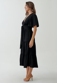 Black tiered midi dress with short flutter sleeves, V-neckline, and a waist tie. Made from lightweight, textured fabric. Stiletto heels.