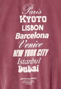 Bordowy T-shirt z tkaniny z białym napisem wymieniającym miasta: Paryż, Kioto, Lizbona, Barcelona, Wenecja, Nowy Jork, Stambuł, Dubaj.