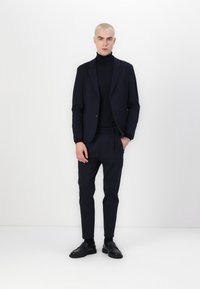 Navy suit with a tailored blazer and tapered trousers, complemented by a fitted black turtleneck and black loafers. Smooth fabric texture.