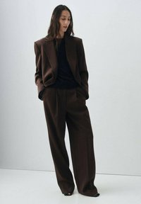 Blazer oversize marron associé à un pantalon marron évasé. Tissu doux et texturé ; design minimaliste ; haut noir élégant ; chaussures ouvertes noires.