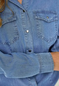 Chemise en denim bleu clair à manches longues, fermeture à boutons, deux poches poitrine et boutons-pression argentés. Texture douce avec un design classique.