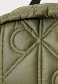 Sac à dos olive quilté avec un motif géométrique, des accents cousus et une texture lisse. Comprend une fermeture éclair en haut.