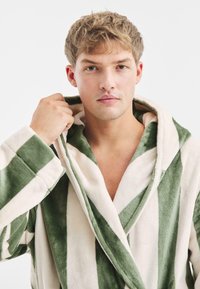 Groene en crèmekleurige gestreepte badjas met een capuchon, gemaakt van zacht, dik materiaal. Beschikt over ruime mouwen en een sjawl-kraag.