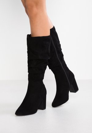 Jambes portant des bottes à talons en daim noir mi-mollet, avec des bouts pointus et une fermeture éclair latérale, posées sur un fond blanc.