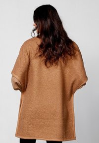 Bruine, gestructureerde oversized buitenjas met korte mouwen, voorzien van een gladde beige afwerking langs de randen en zijopeningen voor meer bewegingsvrijheid.