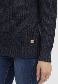 Dark blue knitted sweater with a textured pattern, ribbed cuffs, and a wooden brand tag on the bottom left side.